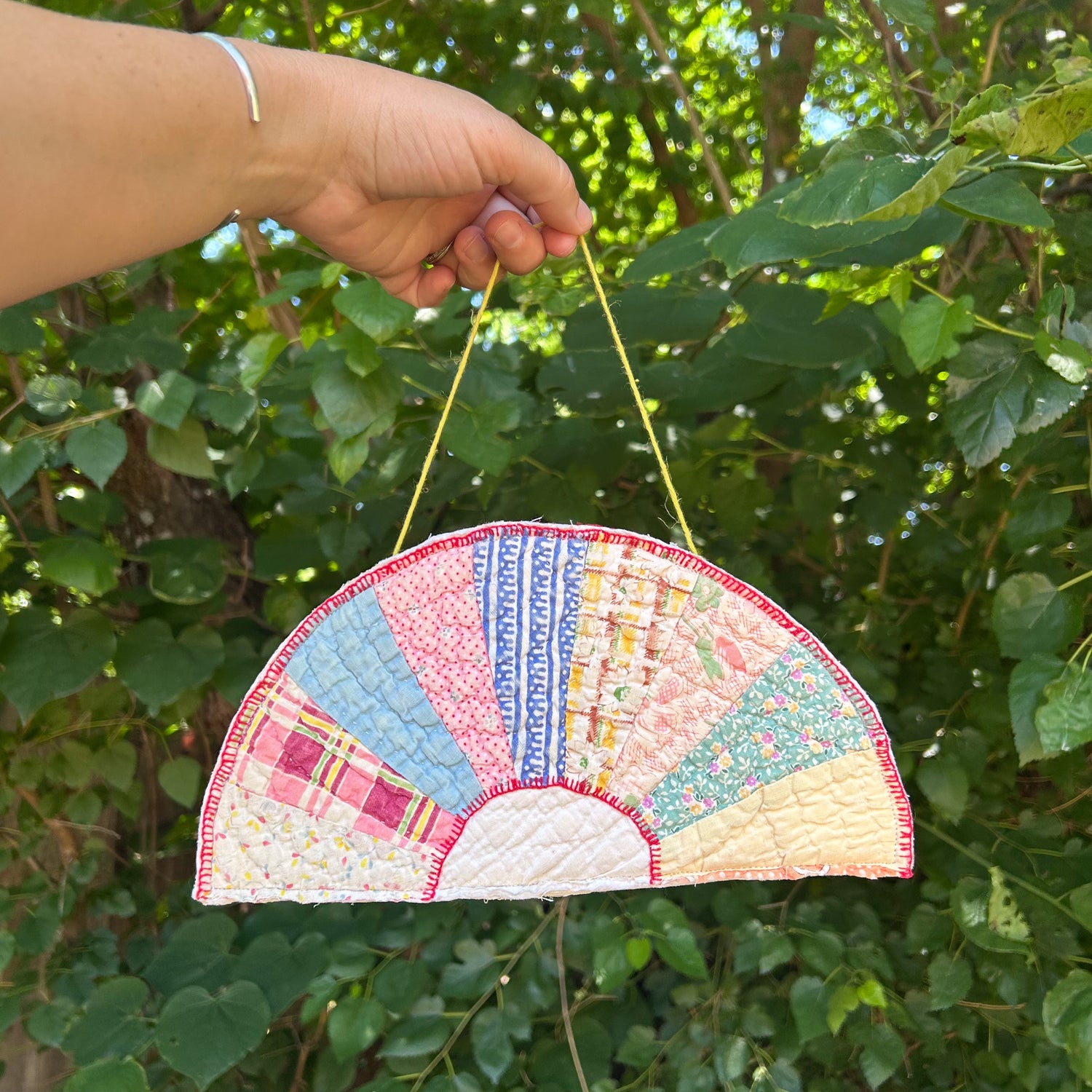 Handmade quilted fan-shaped ornament held by a hand against a green leafy background