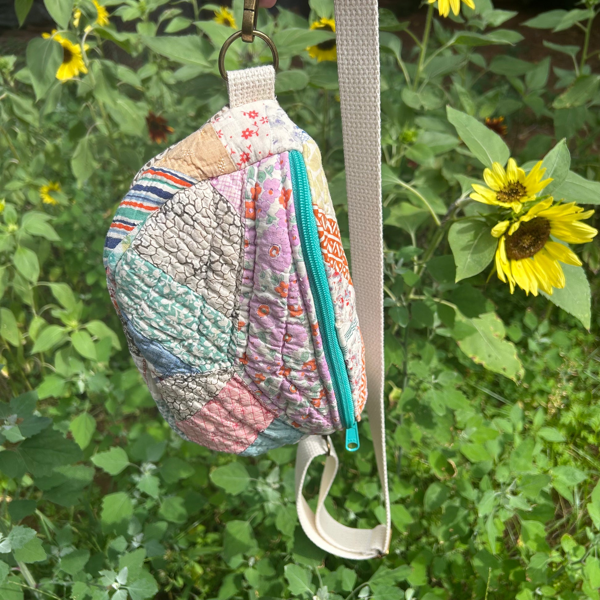 Patchwork-style pouch with a strap, hanging among green foliage and a sunflower.
