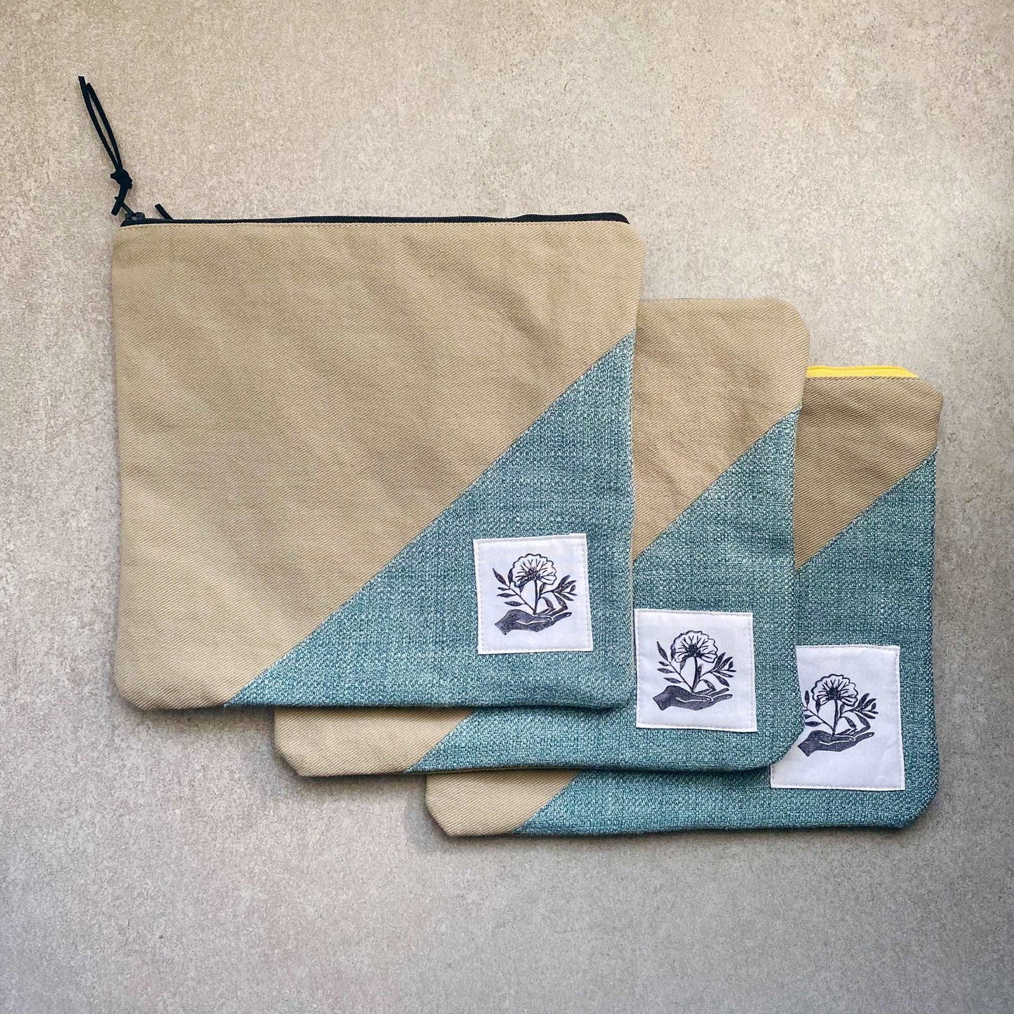 This is a photo of three large canvas zipper pouches that have textured teal fabric panels on them. They are laying on a light gray background.