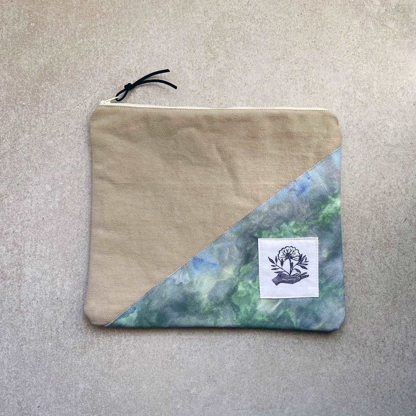 This is a photo of a large canvas zipper pouch featuring a green and blue ice dyed fabric panel. It is laying on a light gray background.