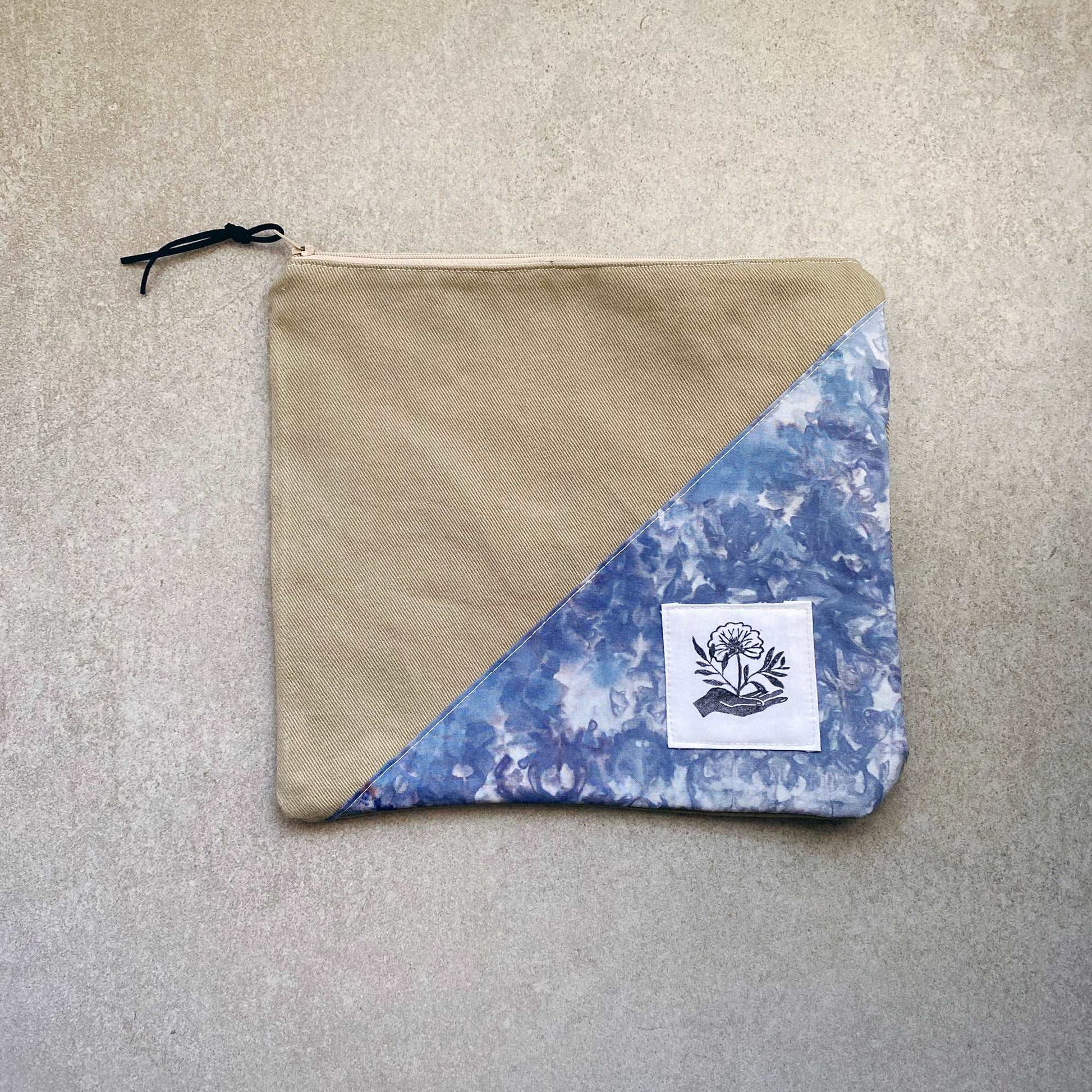 This is a photo of a large canvas zipper bag featuring a blue and white ice dyed fabric panel. It is laying on a light gray background.