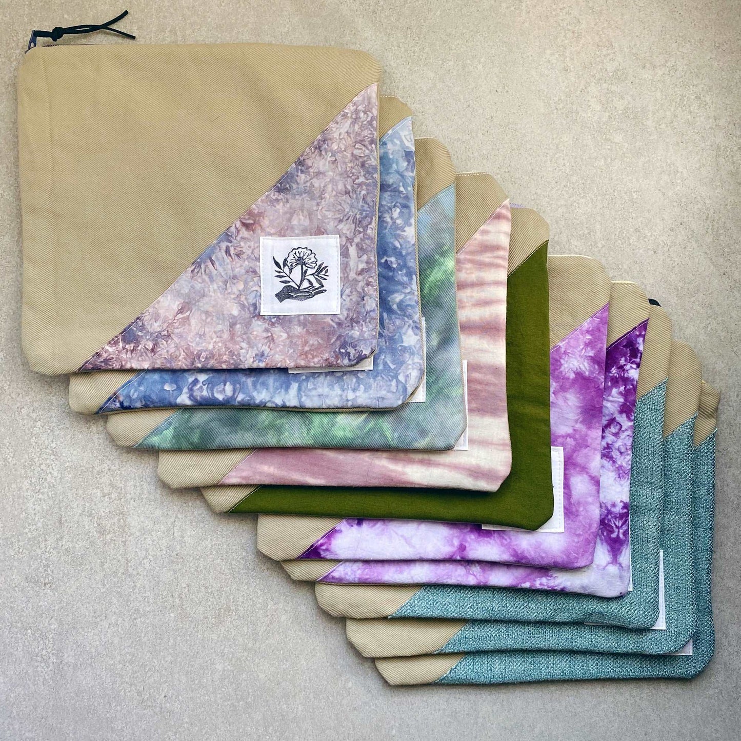 This is a photo of multiple canvas zip bags. Each bag is a little different, with fabric panels made from tie dyed fabric, solid fabric and textured fabric. Each bag includes a handprinted patch made by Sunny Chan of the Etsy shop moonandmountainprint. 