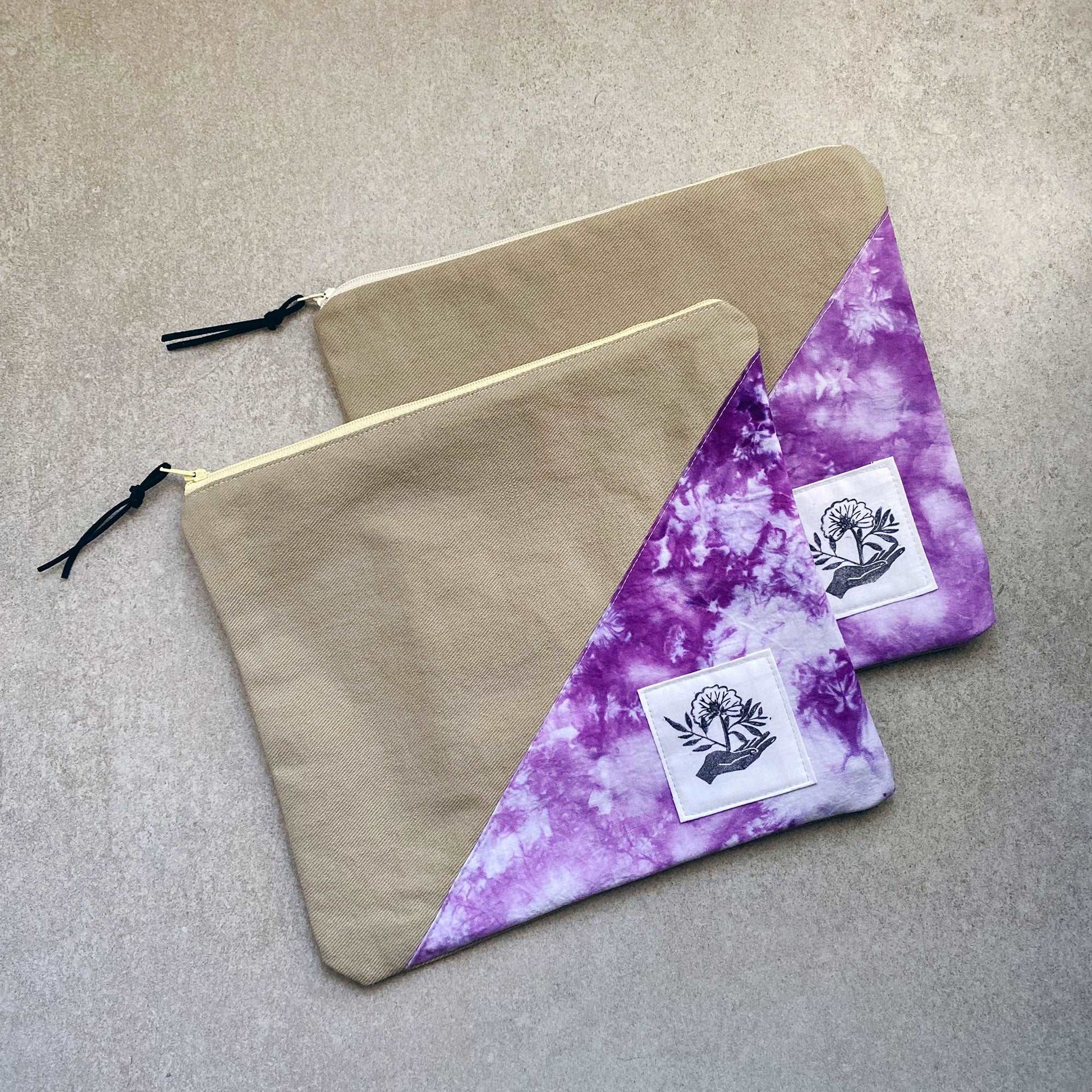 This is a photo of two large canvas zipper bags made with purple and white tie dye fabric panels. They are laying on a light gray background.