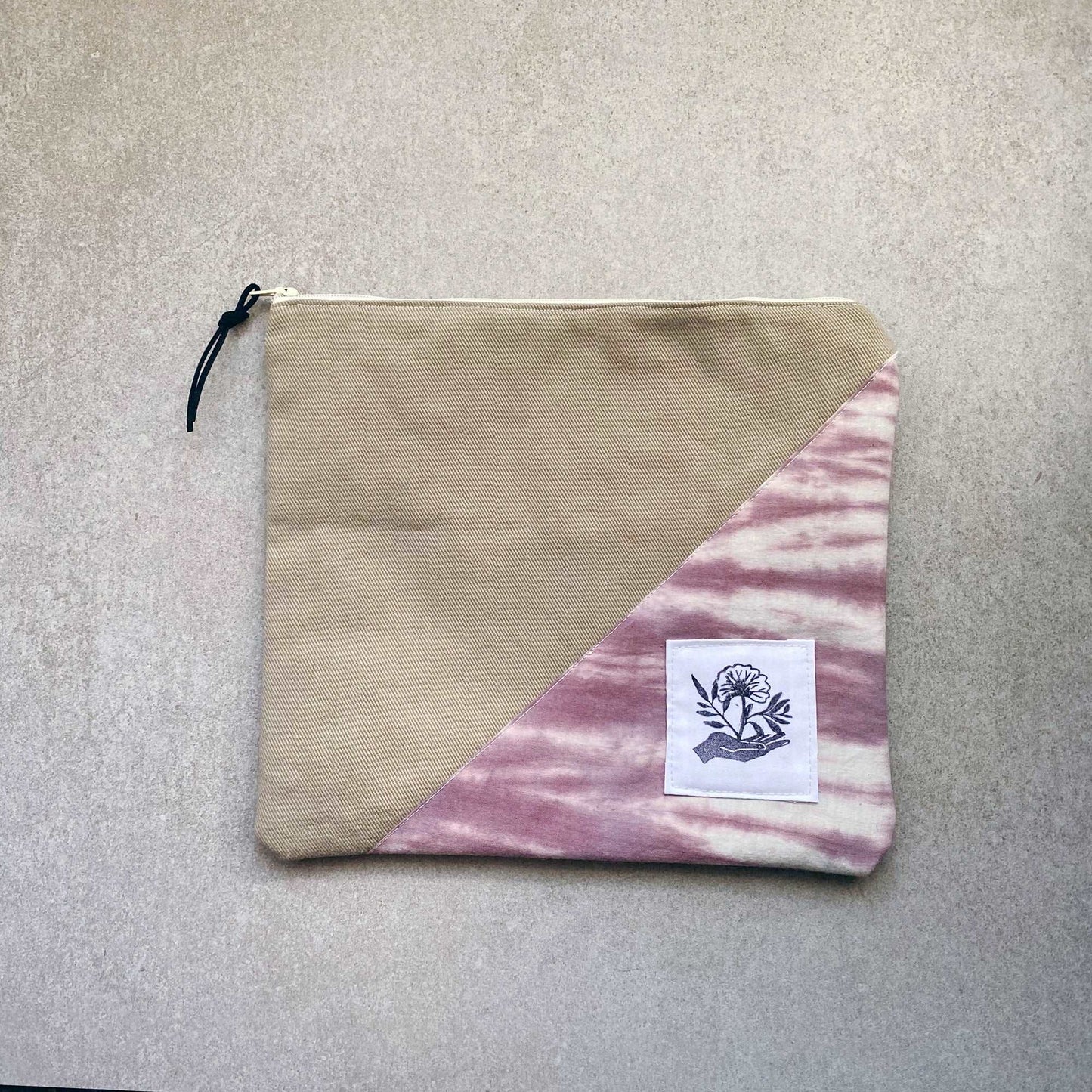This is a photo of a large canvas zipper pouch with a clay colored tie dye fabric panel on it. It is laying on a light gray background.