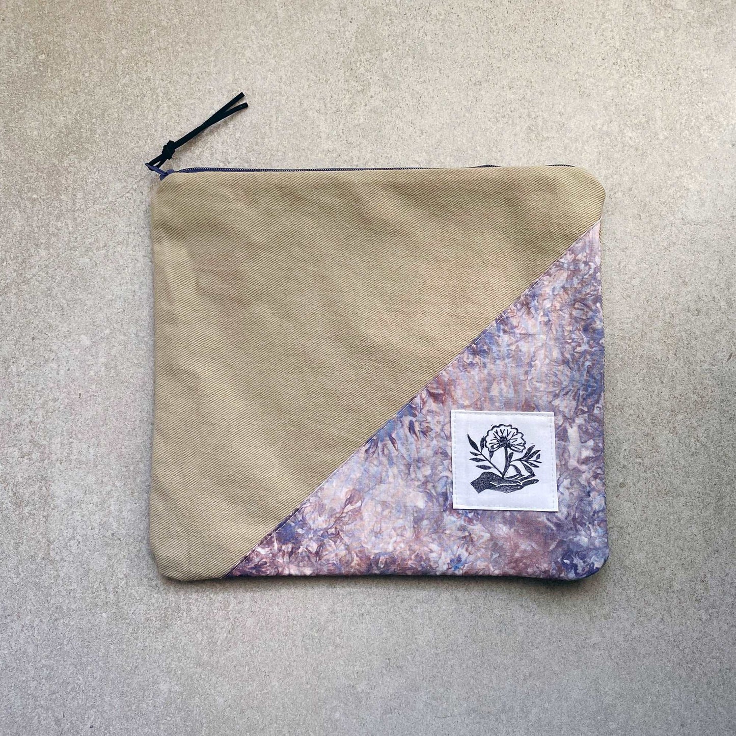 This is a photo of a large canvas zipper bag. It has a fabric panel made with ice dyed fabric that has several colors of purple and blue on it. It is laying on a light gray background.